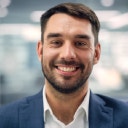 Primo piano di un uomo sorridente con la barba, che indossa una giacca blu e una camicia bianca in un ufficio luminoso.