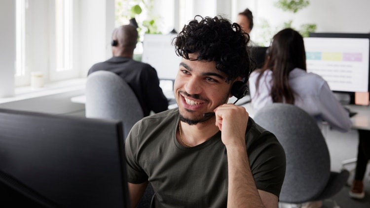 Un giovane operatore con cuffie microfoniche sorride mentre lavora in un ufficio open space luminoso.
