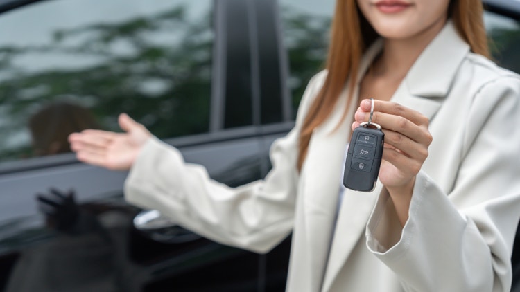 Una donna in giacca bianca porge la chiave elettronica di un&#39;auto scura con un gesto di accoglienza.