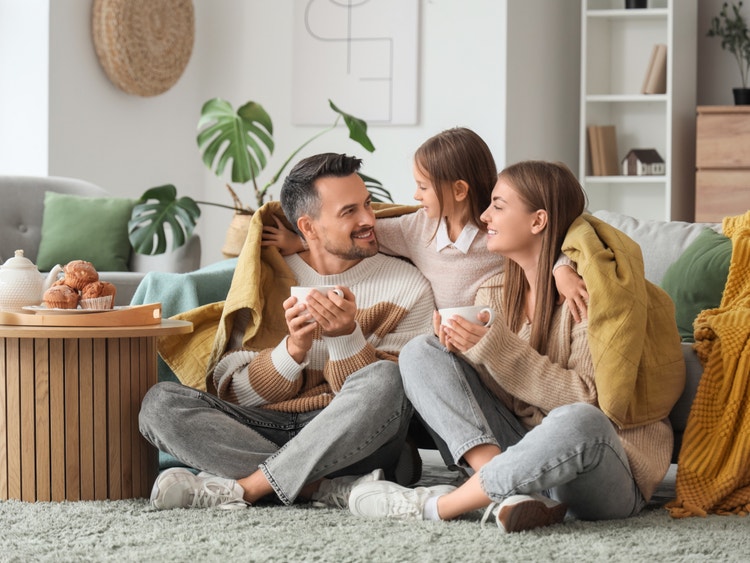 Famiglia felice seduta sul tappeto sotto una coperta gialla, mentre beve tè in un soggiorno moderno e accogliente.