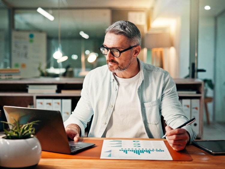 Uomo con occhiali e barba brizzolata lavora al laptop analizzando grafici cartacei in un ufficio moderno e luminoso.