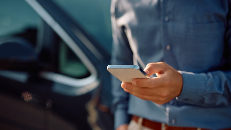 Primo piano delle mani di un uomo che utilizza uno smartphone, con un&#39;automobile scura sfocata sullo sfondo.