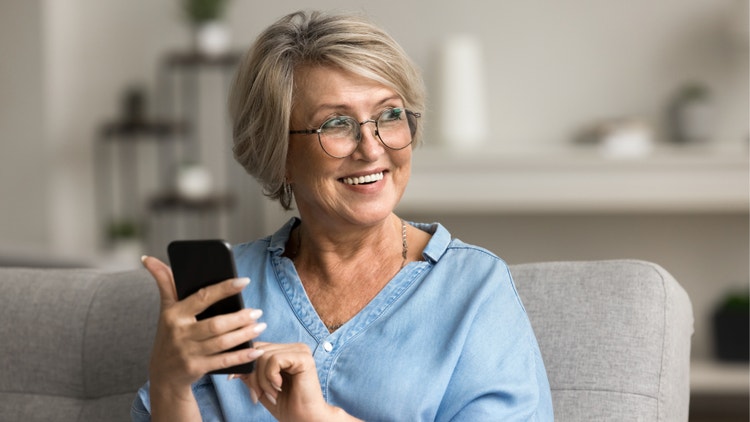 Donna sorridente con occhiali e camicia azzurra mentre usa uno smartphone seduta sul divano di casa.