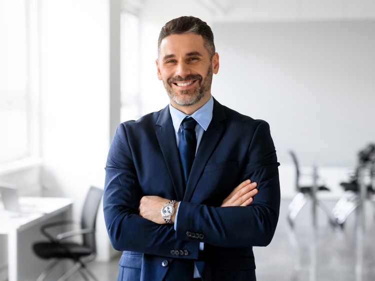 Uomo in giacca e cravatta sorridente con le braccia conserte in un ufficio moderno.