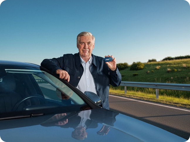 Primo piano di Carlo Ancelotti appoggiato a un&#39;auto scura che mostra il dispositivo Unibox in mano.
